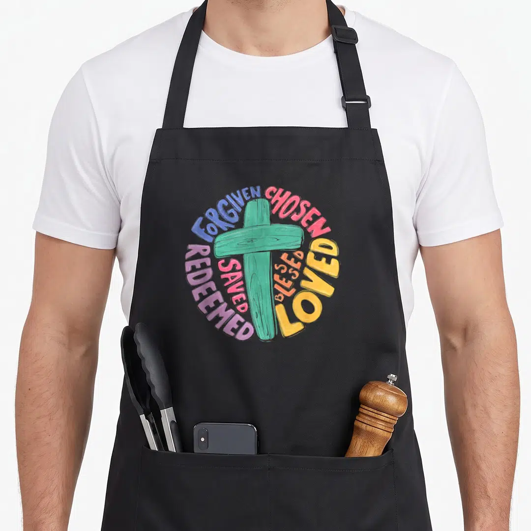 Cross easter apron - Teal cross with circular colorful text reading Forgiven Chosen Loved Blessed Saved Redeemed — shown on a black cotton canvas apron against a white background