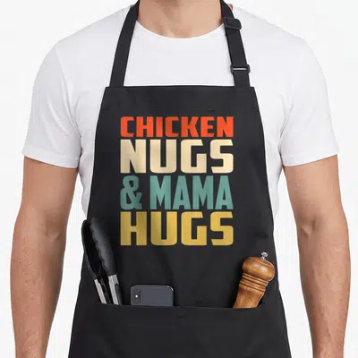 Hug nugget chicken print apron - Stacked bold text reading CHICKEN NUGS & MAMA HUGS in orange beige teal and yellow — shown on a black cotton canvas apron against a white background