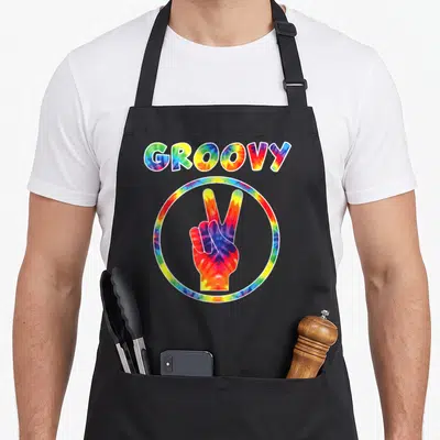 Groovy tie-dye apron - GROOVY text above tie-dye circle containing hand making peace sign in red yellow green blue — shown on a black cotton canvas apron against a white background