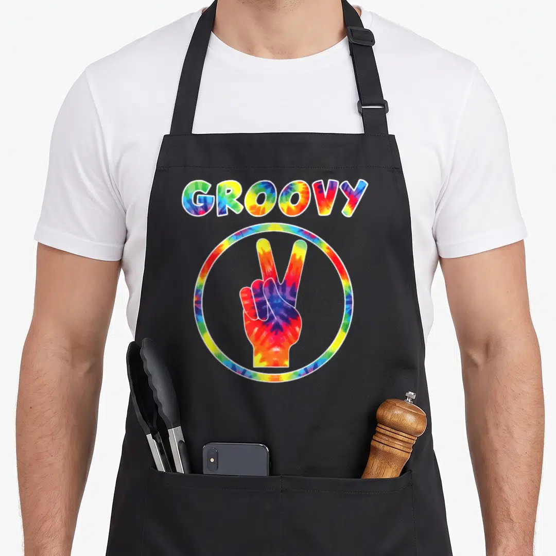 Groovy tie-dye apron - GROOVY text above tie-dye circle containing hand making peace sign in red yellow green blue — shown on a black cotton canvas apron against a white background