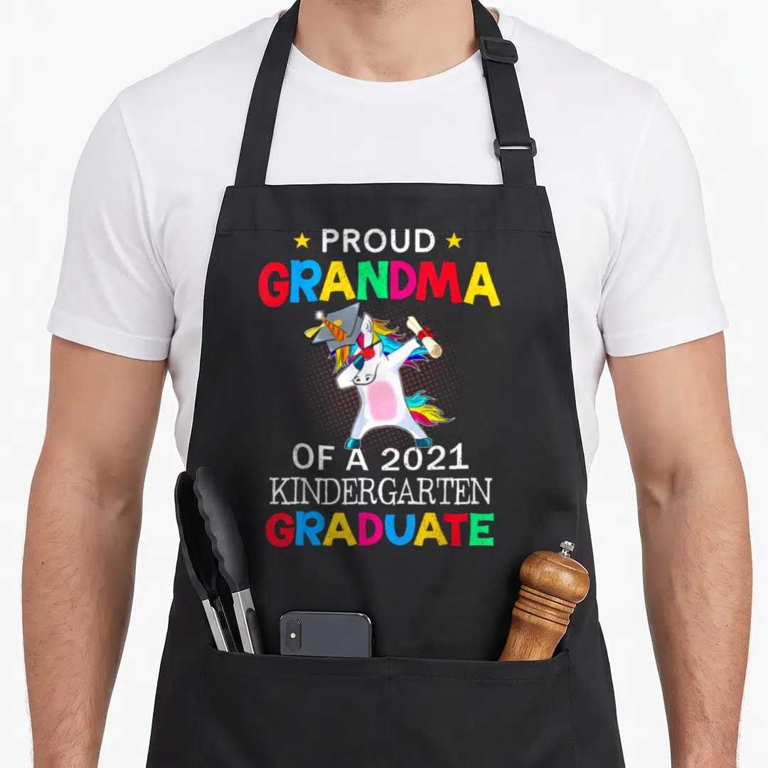 Proud unicorn aprons - Cartoon white unicorn dabbing in cap holding diploma with stacked colorful graduate text — shown on a black cotton canvas apron against a white background