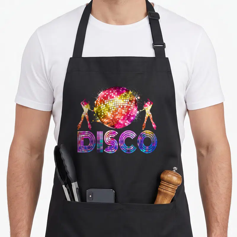 Disco theme 70s apron - Colorful disco ball graphic with two dancers and DISCO text in pink yellow blue purple — shown on a black cotton canvas apron against a white background
