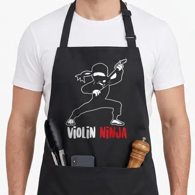 Ninja violin apron - White line-art ninja playing violin above white and red text — shown on a black cotton canvas apron against a white background