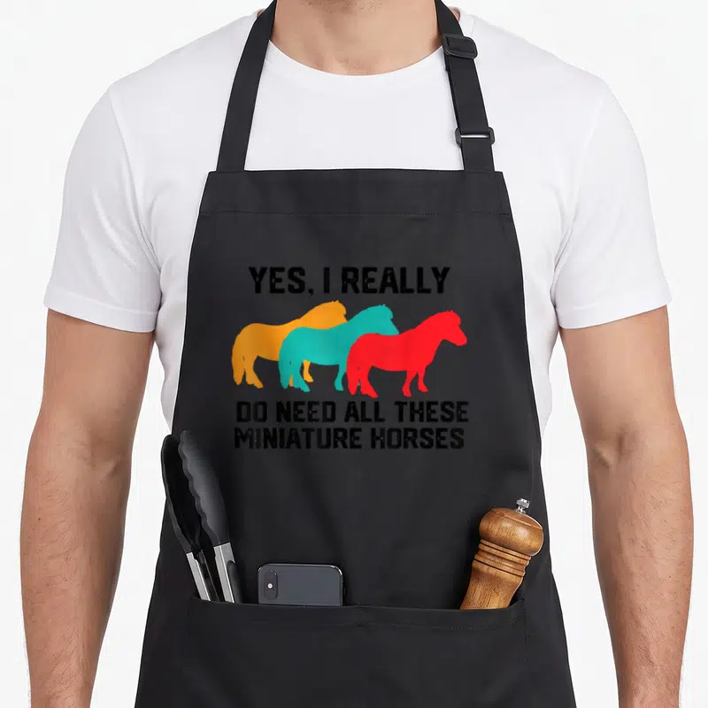 Horse do i need an apron - Three overlapping orange teal and red miniature horse silhouettes between distressed black — shown on a black cotton canvas apron against a white background