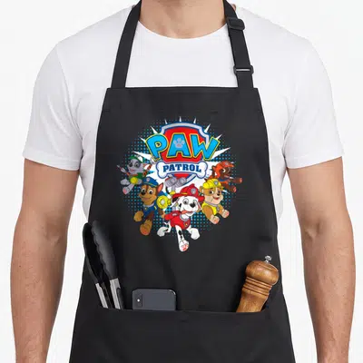 Funny paw apron - PAW PATROL shield with five puppies and binoculars in blue red yellow — shown on a black cotton canvas apron against a white background