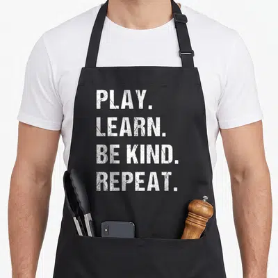 Play learning apron - White distressed text stacked vertically reading PLAY LEARN BE KIND REPEAT — shown on a black cotton canvas apron against a white background
