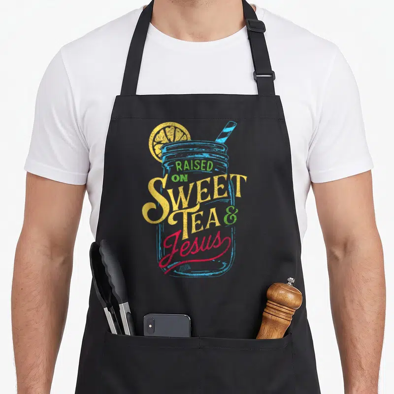 Raised southern aprons - Retro mason jar with lemon slice and straw. Stacked text Raised On Sweet Tea & Jesus — shown on a black cotton canvas apron against a white background