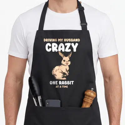 Rabbit animal driving aprons - Sitting cartoon rabbit with text driving my husband crazy one rabbit at a time — shown on a black cotton canvas apron against a white background