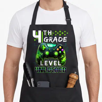 4th level apron - Game controller with 4th grade level unlocked text and green plus signs — shown on a black cotton canvas apron against a white background