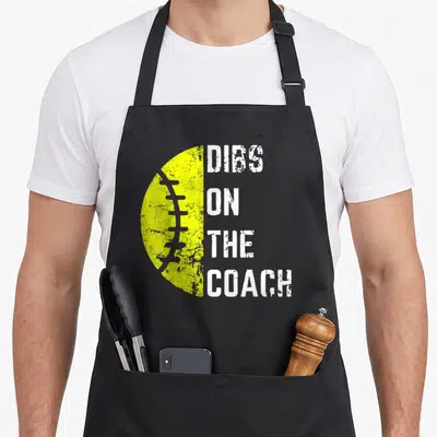 Softball wife apron - Yellow half-football next to stacked DIBS ON THE COACH text in distressed white font — shown on a black cotton canvas apron against a white background