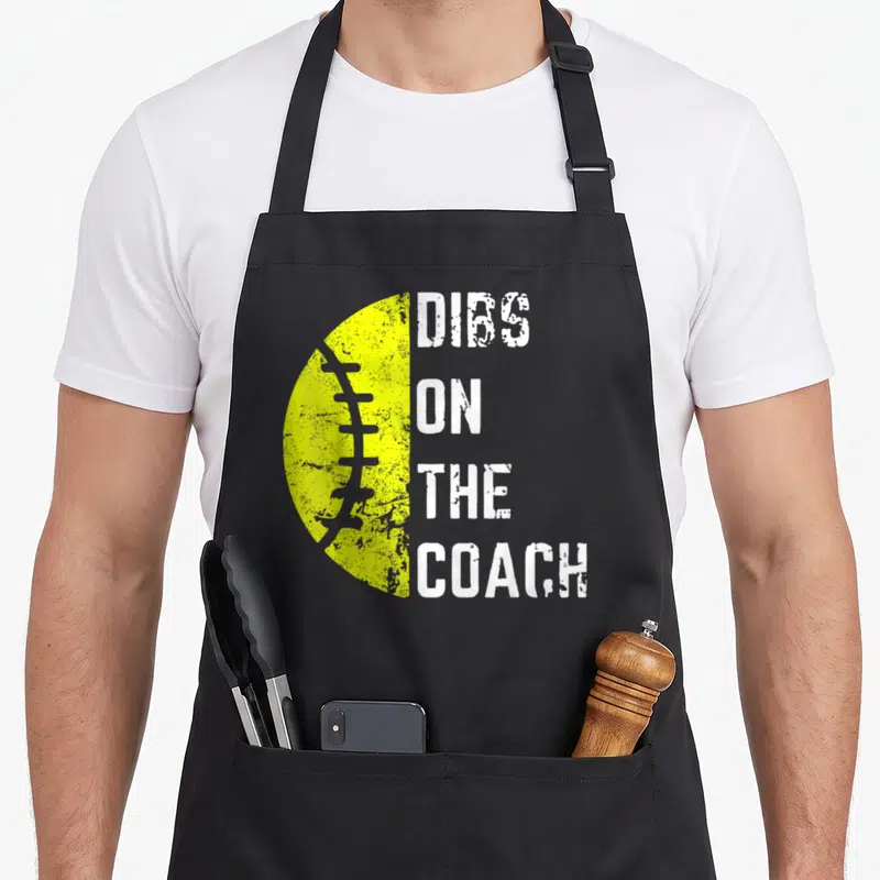 Softball wife apron - Yellow half-football next to stacked DIBS ON THE COACH text in distressed white font — shown on a black cotton canvas apron against a white background