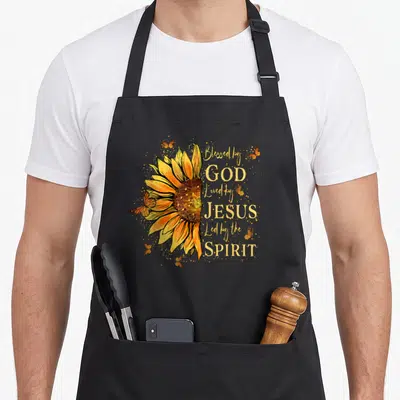 Blessed led apron - Sunflower with yellow orange petals and text Blessed by God Loved by Jesus Led by the Spirit — shown on a black cotton canvas apron against a white background