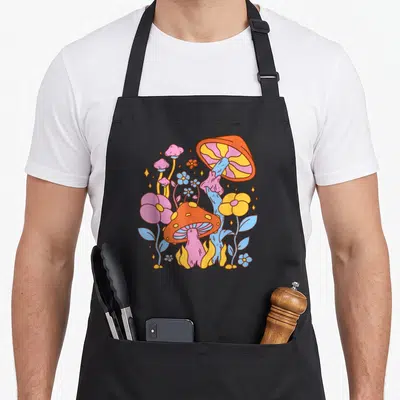 Hippy cottage apron - Cartoon cluster of orange pink blue and yellow mushrooms and flowers — shown on a black cotton canvas apron against a white background