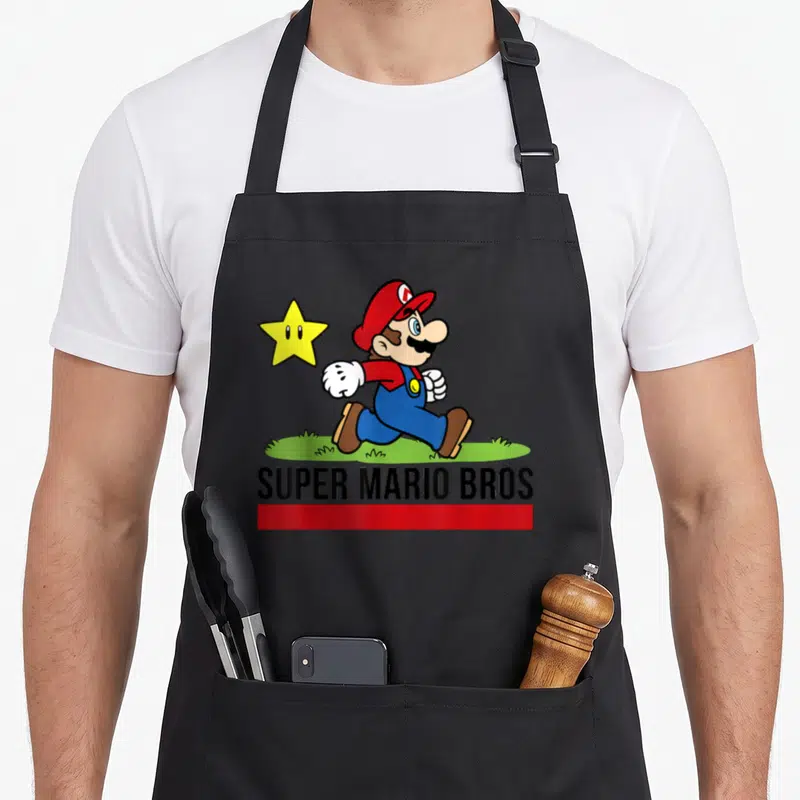 Star walking apron - Mario running on grass with yellow star and SUPER MARIO BROS text — shown on a black cotton canvas apron against a white background