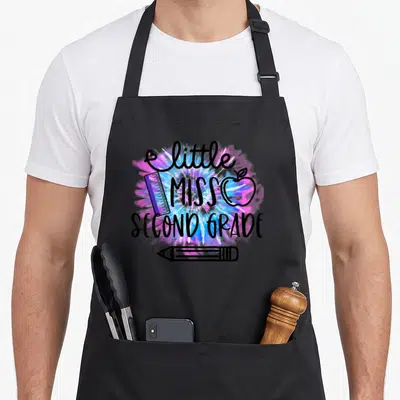 Miss 2nd tie-dye apron - little MISS SECOND GRADE text with ruler apple pencil on blue pink purple tie-dye splash — shown on a black cotton canvas apron against a white background