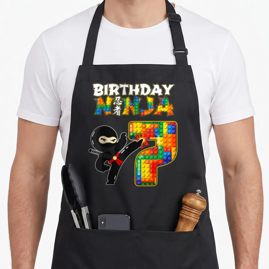 Ninja building apron - Cartoon ninja kicking colorful brick number 7 with BIRTHDAY NINJA text — shown on a black cotton canvas apron against a white background