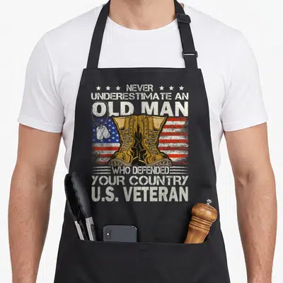 Patriot father day apron - Boots over flag with dog tags and veteran text in distressed white, tan, red, and blue — shown on a black cotton canvas apron against a white background