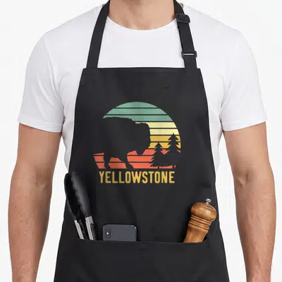Travel buffalo apron - Grey bison silhouette against striped yellow orange red and green sun with YELLOWSTONE text below — shown on a black cotton canvas apron against a white background