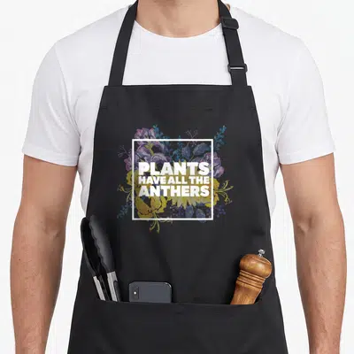 Anther plant apron - White text PLANTS HAVE ALL THE ANTHERS over purple yellow blue watercolor flowers in white square — shown on a black cotton canvas apron against a white background
