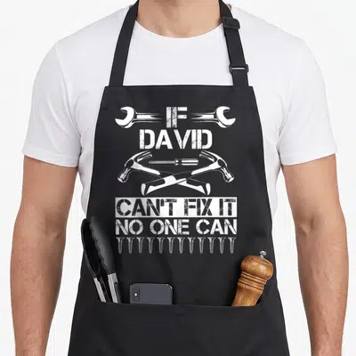 David father's day apron - White distressed text If David Can't Fix It No One Can with crossed hammers and screws — shown on a black cotton canvas apron against a white background
