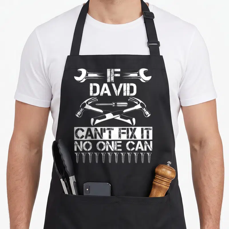David father's day apron - White distressed text If David Can't Fix It No One Can with crossed hammers and screws — shown on a black cotton canvas apron against a white background