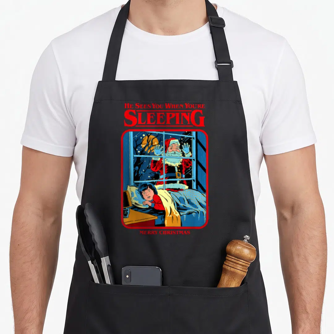 Steven santa claus apron - Red text He Sees You When You're Sleeping above cartoon Santa peeking through window — shown on a black cotton canvas apron against a white background
