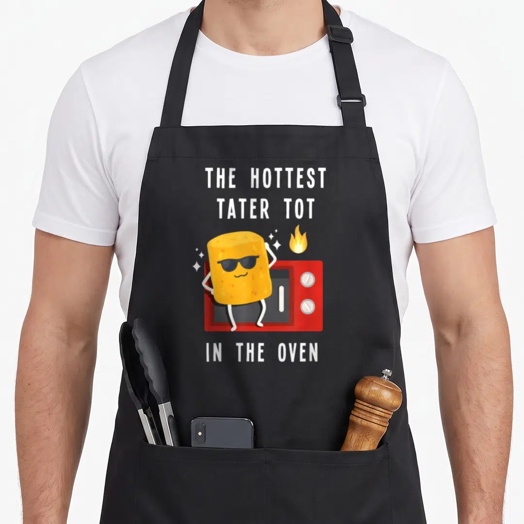 Hottest oven apron - Cartoon tater tot in sunglasses standing before red oven with white text — shown on a black cotton canvas apron against a white background