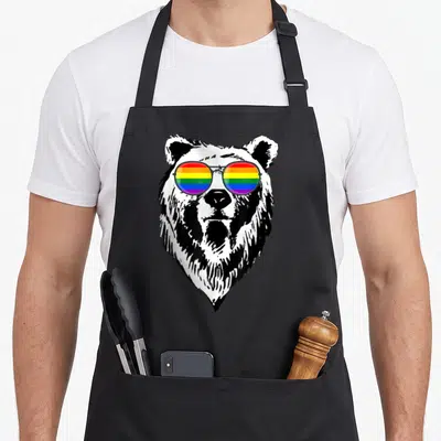 Bear gay pride apron - Cartoon bear head with rainbow-striped sunglasses printed on cotton canvas apron — shown on a black cotton canvas apron against a white background