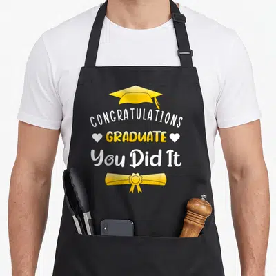Graduate funny graduation apron - Yellow cap, white hearts, diploma scroll, and congratulatory text on black background — shown on a black cotton canvas apron against a white background