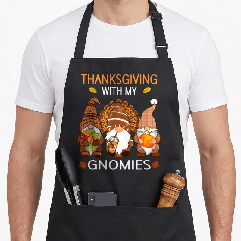 Fall thanksgiving apron - Three cartoon gnomes holding autumn leaves, a drink, and a pumpkin under THANKSGIVING WITH MY — shown on a black cotton canvas apron against a white background