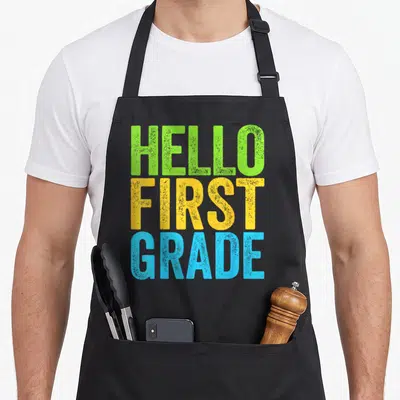 Grade hello apron - Stacked distressed text HELLO FIRST GRADE in green yellow and blue — shown on a black cotton canvas apron against a white background