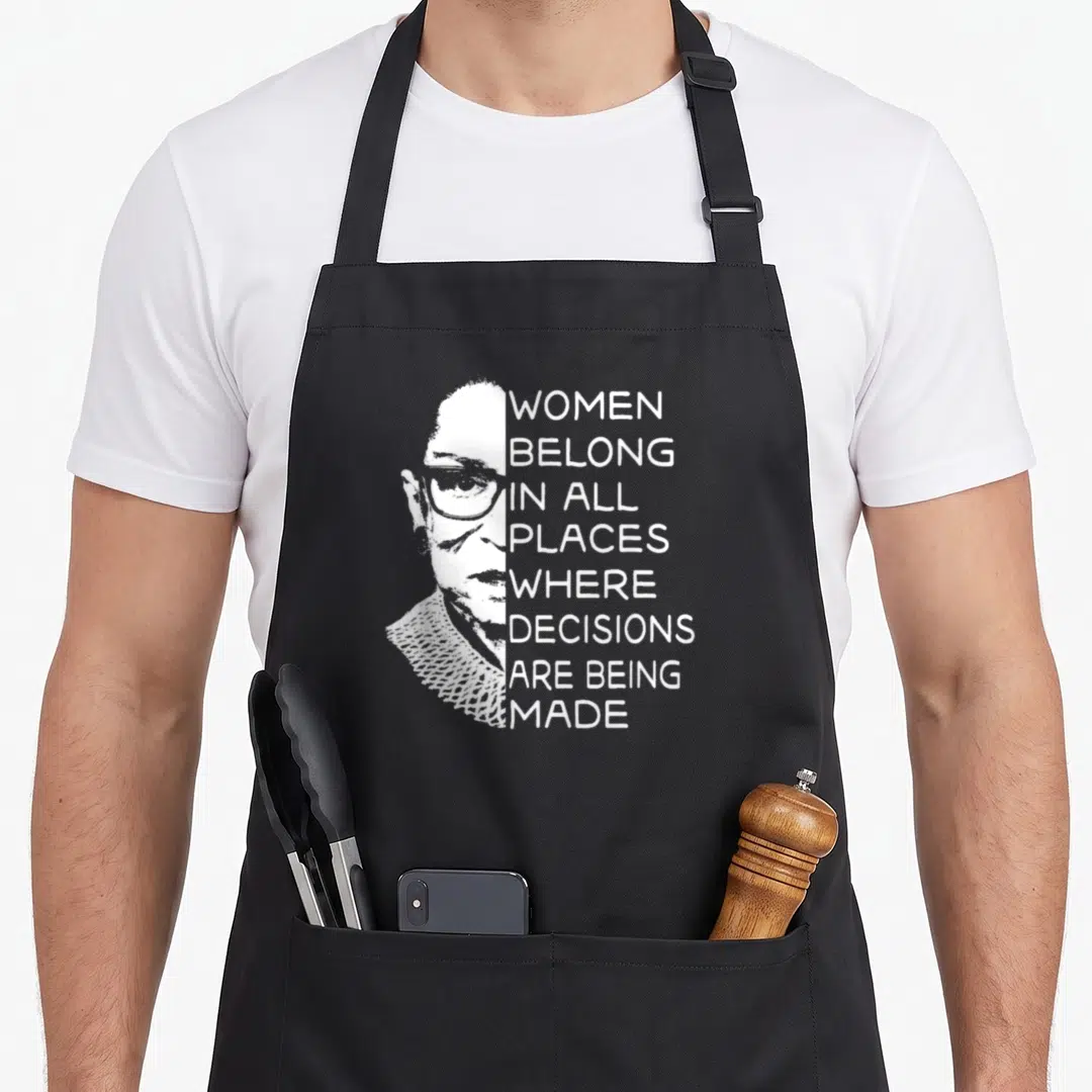 Notorious roller lover apron - White portrait of a woman next to stacked text WOMEN BELONG IN ALL PLACES WHERE — shown on a black cotton canvas apron against a white background