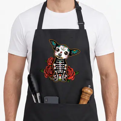 Calavera colorful apron - Cartoon sugar skull chihuahua sitting among three red roses and green leaves — shown on a black cotton canvas apron against a white background