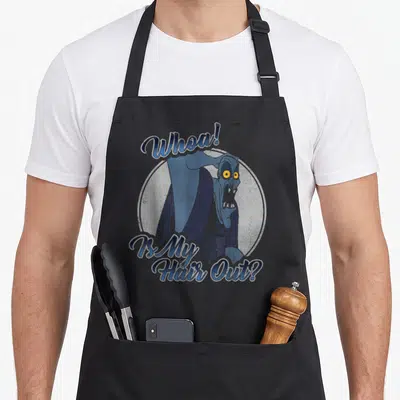 Hercule hair apron - Blue cartoon character with yellow eyes and text Whoa Is My Hair Out inside grey circle — shown on a black cotton canvas apron against a white background