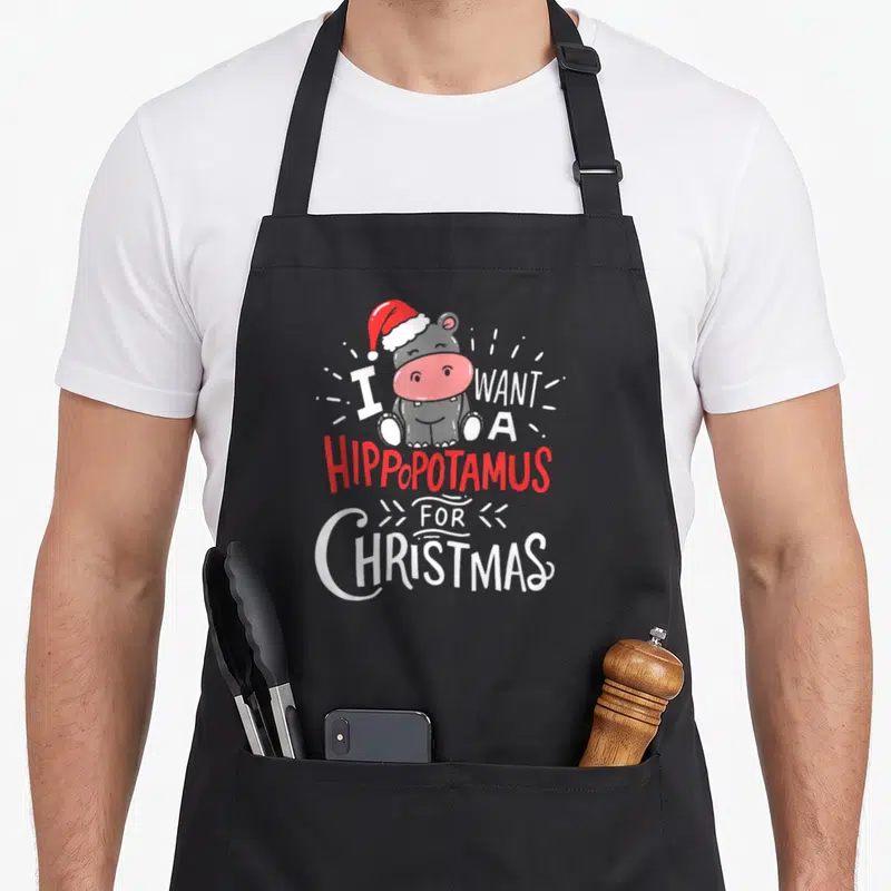 Xmas hippo christmas apron - Cartoon grey hippo in red hat with text I WANT A HIPPOPOTAMUS FOR CHRISTMAS — shown on a black cotton canvas apron against a white background