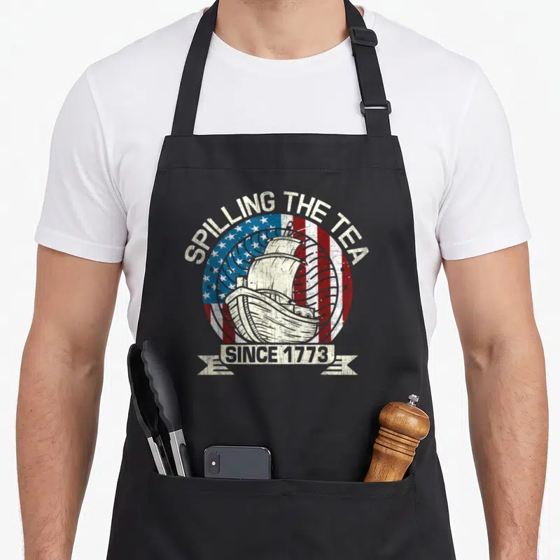 Patriotic tea party aprons - Distressed ship and flag circle with SPILLING THE TEA SINCE 1773 text — shown on a black cotton canvas apron against a white background