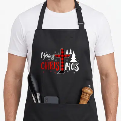 Christian buffalo plaid christmas apron - Red black plaid cross splitting Christmas text with white trees and snowflakes — shown on a black cotton canvas apron against a white background