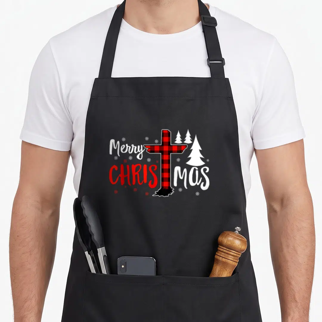 Christian buffalo plaid christmas apron - Red black plaid cross splitting Christmas text with white trees and snowflakes — shown on a black cotton canvas apron against a white background