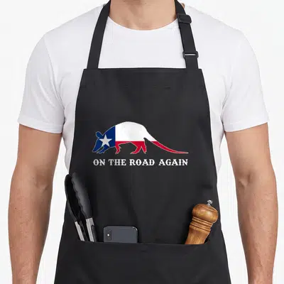 Road texas flag apron - Armadillo silhouette filled with blue white red Texas flag pattern above white text — shown on a black cotton canvas apron against a white background