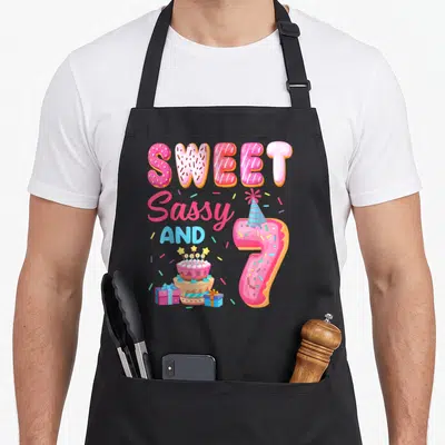 Theme sassy apron - Pink donut text reading Sweet Sassy and 7 with cake and party hat — shown on a black cotton canvas apron against a white background