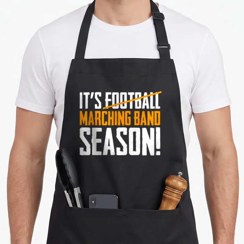 Band football apron - Distressed white and orange text reading IT'S FOOTBALL MARCHING BAND SEASON with orange — shown on a black cotton canvas apron against a white background