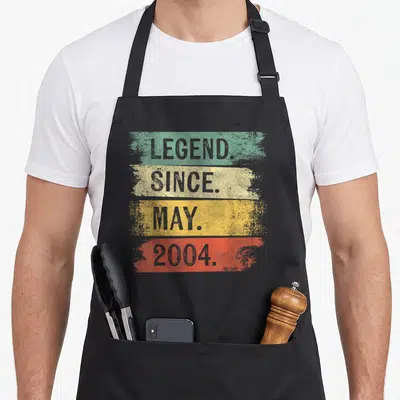 Legend may birthday apron - Four colored stripes with distressed grey text reading LEGEND SINCE MAY 2004 — shown on a black cotton canvas apron against a white background