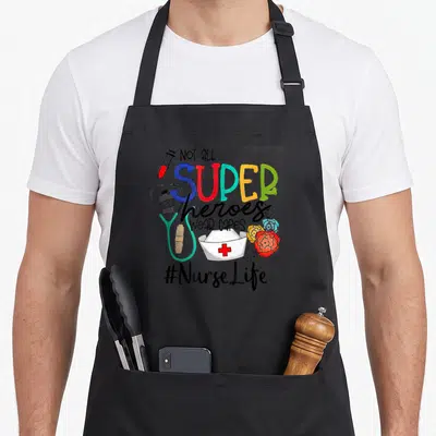 Nurse mother's day aprons - Stacked text not all super heroes wear capes with cartoon syringe stethoscope nurse hat — shown on a black cotton canvas apron against a white background