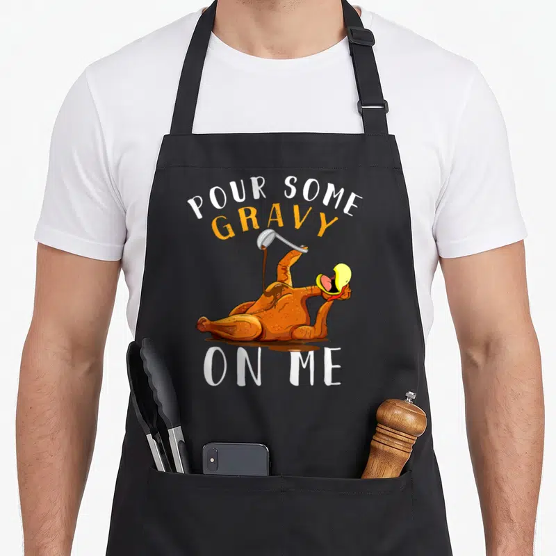 Pour thanksgiving day aprons - Reclining cartoon turkey pouring gravy with text POUR SOME GRAVY ON ME in orange — shown on a black cotton canvas apron against a white background