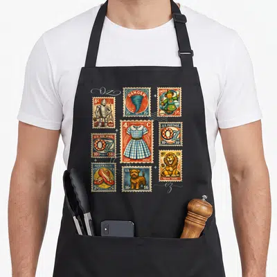 Post wicked apron - Nine vintage Wizard of Oz stamps including Tin Man, Tornado, and Ruby Slippers in red blue yellow — shown on a black cotton canvas apron against a white background