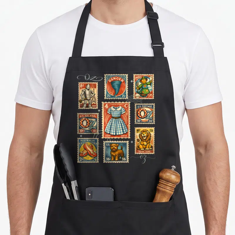Post wicked apron - Nine vintage Wizard of Oz stamps including Tin Man, Tornado, and Ruby Slippers in red blue yellow — shown on a black cotton canvas apron against a white background