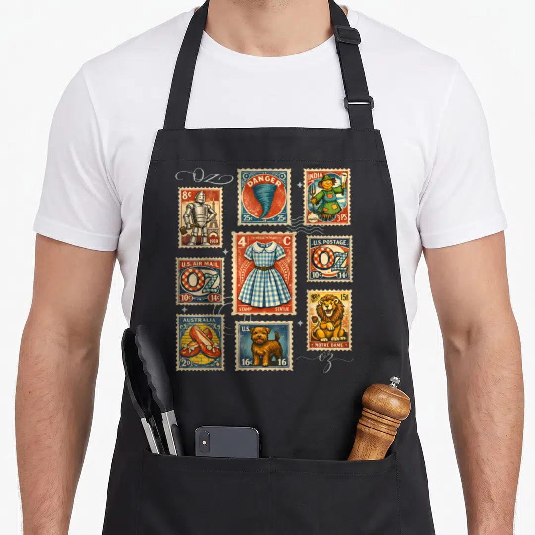 Post wicked apron - Nine vintage Wizard of Oz stamps including Tin Man, Tornado, and Ruby Slippers in red blue yellow — shown on a black cotton canvas apron against a white background
