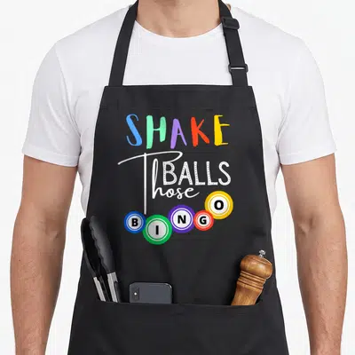 Those bingo apron - SHAKE Balls those text with five colorful BINGO balls in arc arrangement — shown on a black cotton canvas apron against a white background