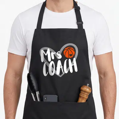 Basketball coach wife apron - Mrs COACH text over white heart outline with orange basketball containing small heart — shown on a black cotton canvas apron against a white background