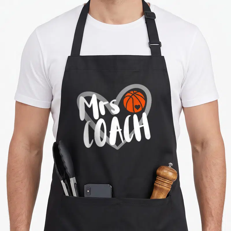 Basketball coach wife apron - Mrs COACH text over white heart outline with orange basketball containing small heart — shown on a black cotton canvas apron against a white background